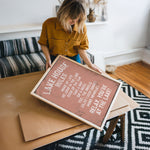 A woman in a yellow shirt is holding a framed sign with text on it, standing in a room with a patterned rug and a couch.