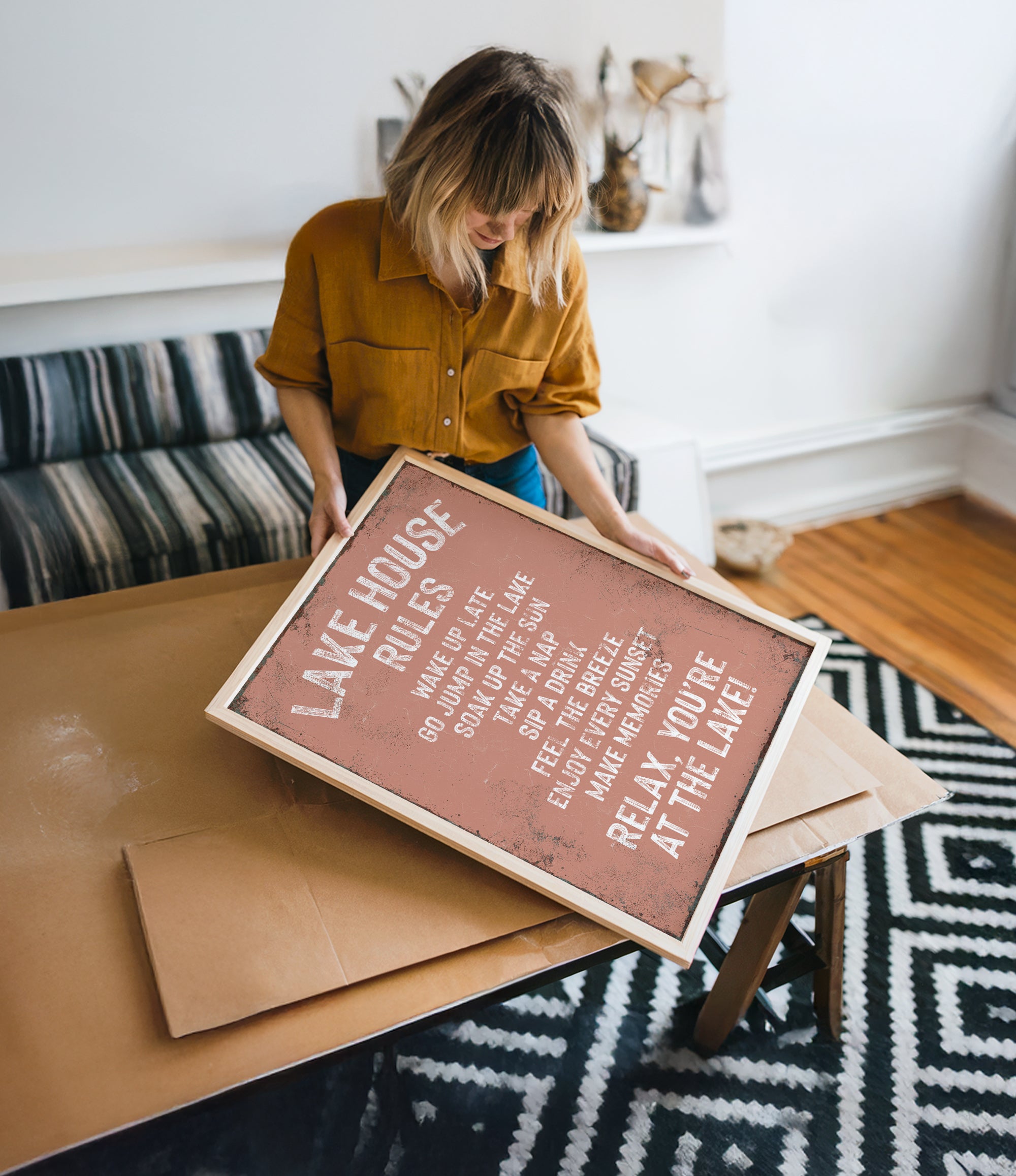 A woman in a yellow shirt is holding a framed sign with text on it, standing in a room with a patterned rug and a couch.
