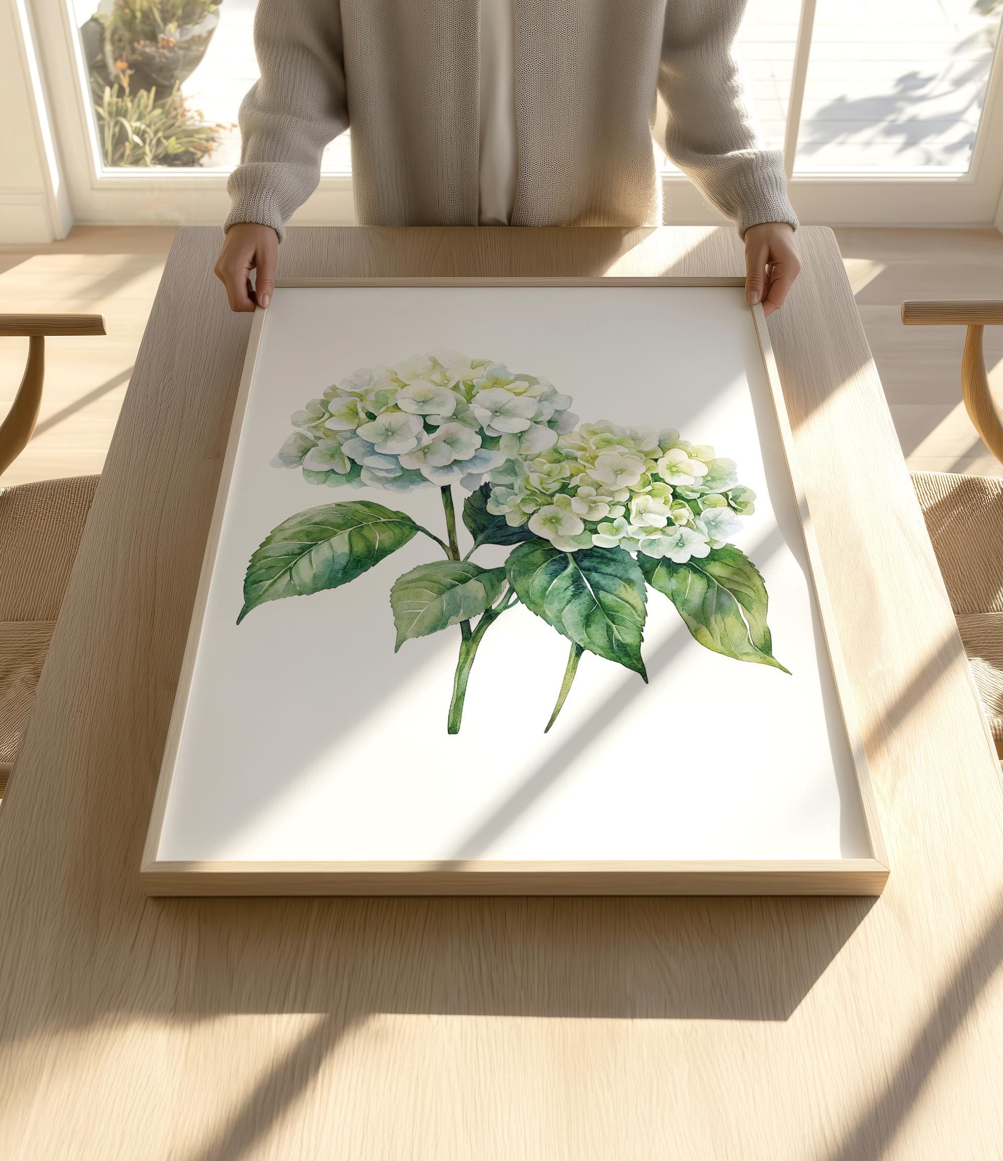 A person is holding up a framed painting of two white hydrangea flowers with green leaves on a wooden table.