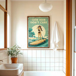 a bathroom with a framed vintage poster of a woman paddleboarding on a lake, a towel hanging on the wall, and a potted plant on the floor.