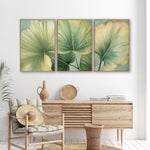 a living room with a wooden dresser, a woven basket, and a woven chair. Above the dresser, there are three framed prints of large green leaves on a white wall.