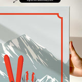 a hand holding a framed print of a mountain landscape with skis and ski poles.