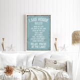 a cozy living room with a white couch, a framed sign on the wall that reads "Lake House Rules", and a vase of flowers on a coffee table.