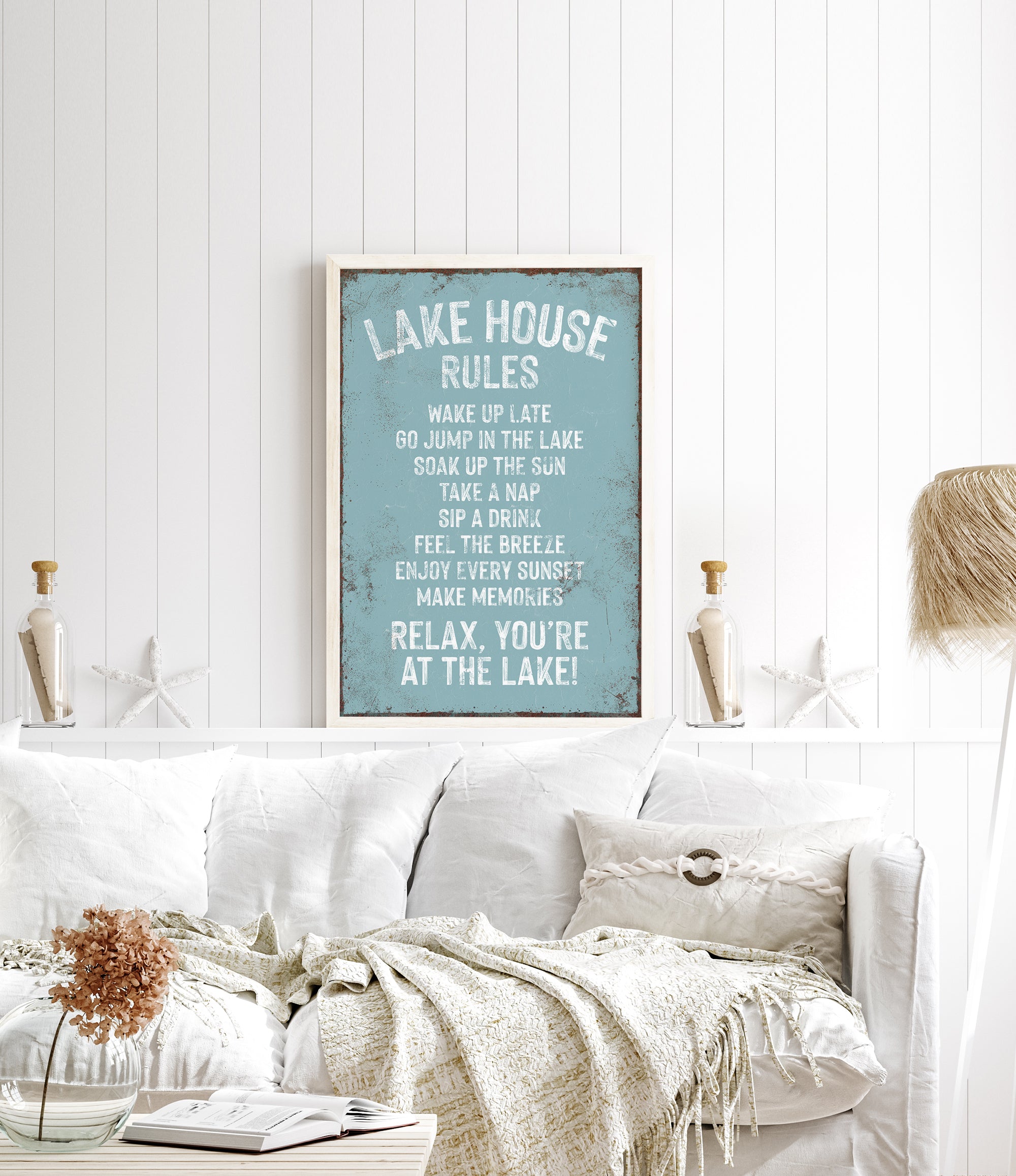 a cozy living room with a white couch, a framed sign on the wall that reads "Lake House Rules", and a vase of flowers on a coffee table.