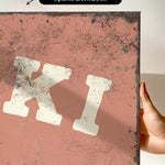 a hand holding a piece of artwork with a distressed, aged appearance. The artwork has a reddish-brown background with splatters and a white, bold letter "K" prominently displayed.