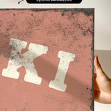 a hand holding a piece of artwork with a distressed, aged appearance. The artwork has a reddish-brown background with splatters and a white, bold letter "K" prominently displayed.