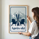 A woman is standing in front of a framed poster that features a mountain landscape with two Adirondack chairs and skis, along with the text "Apres-ski".