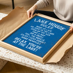 A person is holding a framed sign with a blue background and white text, which appears to be a list of rules or instructions for a lake house.