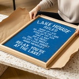 A person is holding a framed sign with a blue background and white text, which appears to be a list of rules or instructions for a lake house.