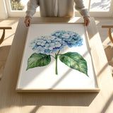A person is holding up a framed painting of a blue hydrangea flower on a wooden table.