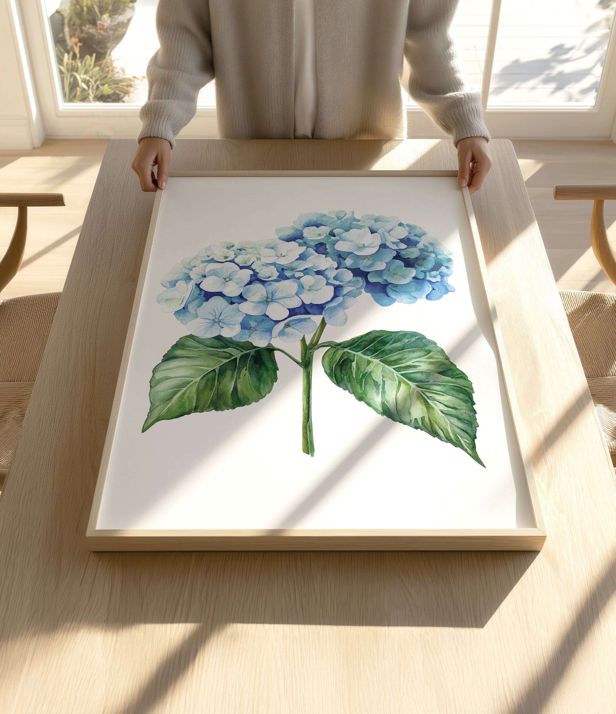 A person is holding up a framed painting of a blue hydrangea flower on a wooden table.