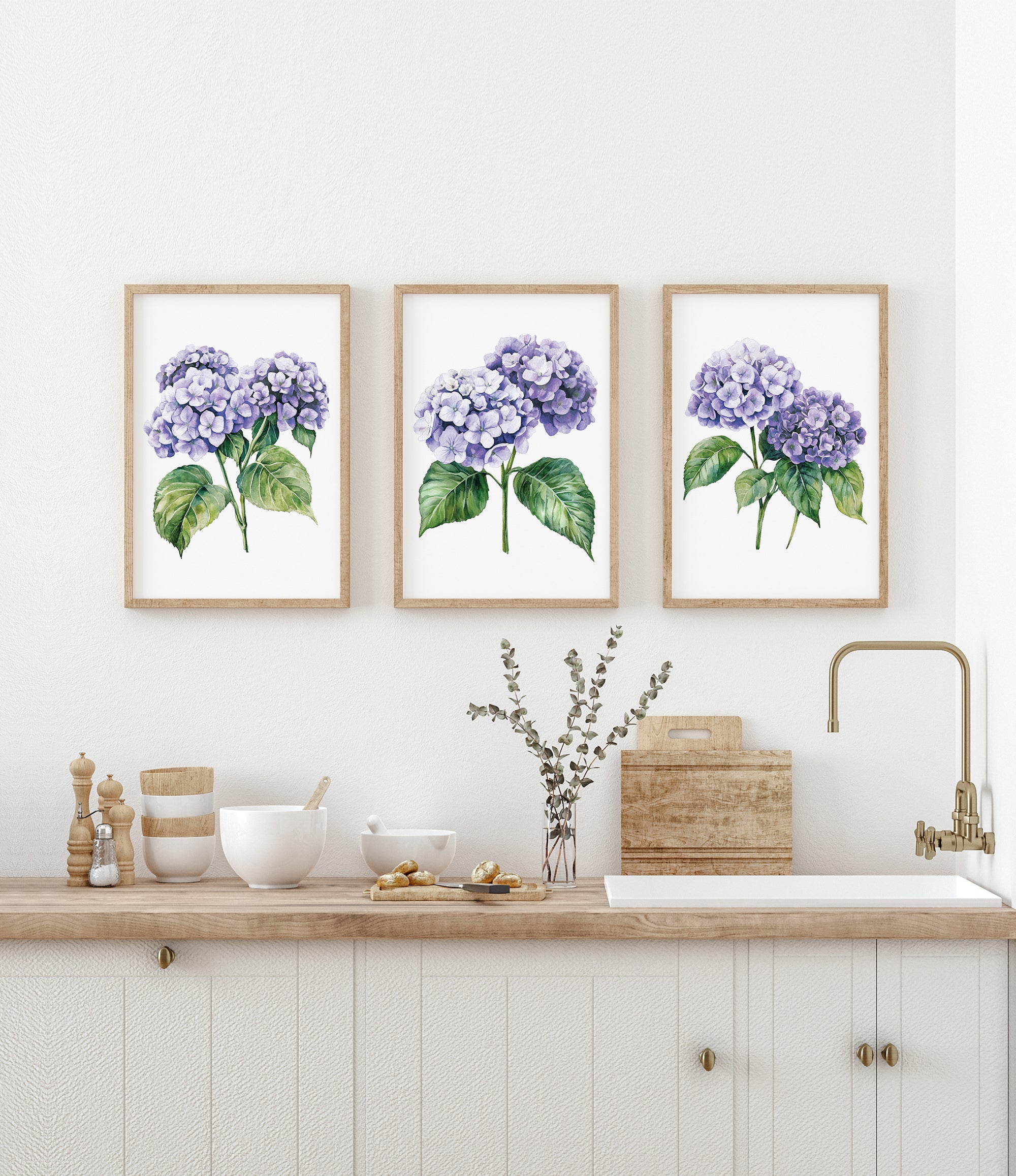 a kitchen counter with three framed watercolor paintings of purple hydrangeas hanging above it. The paintings are arranged in a row, creating a visually appealing display. The kitchen counter also has various items such as bowls, a vase with flowers, and a sink.