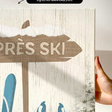 A hand holds the Cerulean Après Ski Signpost Wall Art, a rustic vintage ski sign canvas with blue ski gear—ideal mountain lodge decor for après ski artwork enthusiasts.