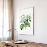 a wooden desk with a lamp, a framed watercolor painting of green hydrangeas, and a window with sheer curtains.