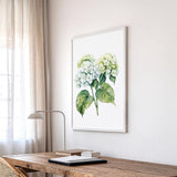 a wooden desk with a lamp, a framed watercolor painting of green hydrangeas, and a window with sheer curtains.