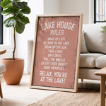 a framed sign with a list of rules for a lake house, placed in a living room with a potted plant and a couch.
