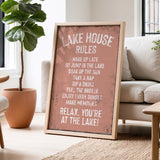 a framed sign with a list of rules for a lake house, placed in a living room with a potted plant and a couch.