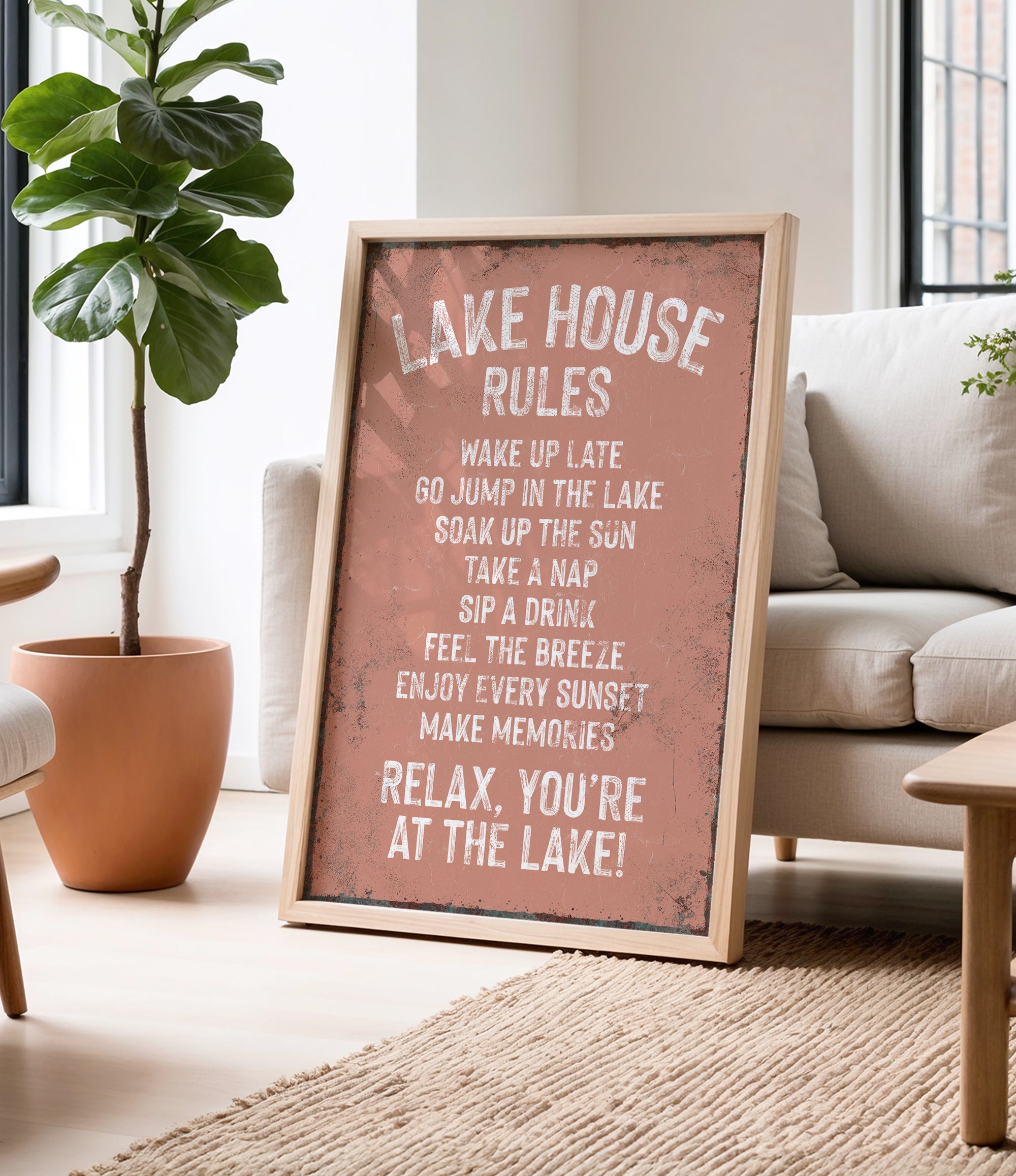 a framed sign with a list of rules for a lake house, placed in a living room with a potted plant and a couch.