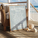 a beach house sign with a beach scene in the background, along with a wicker chair and some seashells on a wooden deck.