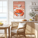 a kitchen with a wicker chair, a wooden table, and a framed painting of a crab on a white and orange striped background. The kitchen has white cabinets and a window overlooking the ocean.