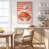 a kitchen with a wicker chair, a wooden table, and a framed painting of a crab on a white and orange striped background. The kitchen has white cabinets and a window overlooking the ocean.