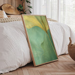 a large framed print of a green leaf with yellow lines, placed on a wooden floor in a bedroom setting.