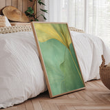 a large framed print of a green leaf with yellow lines, placed on a wooden floor in a bedroom setting.