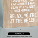 The image is a sign with text that reads "KEEP THE SAND OUTSIDE, HANG UP YOUR TOWEL, HAVE ANOTHER DRINK, ENJOY EVERY SUNSET, MAKE MEMORIES, RELAX, YOU'RE AT THE BEACH!" The sign is made of wood and has a black arrow pointing to it. The sign is described as "ALUMINUM print" and "waterproof, perfect for outdoor use - hanging hardware included".