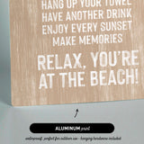 The image is a sign with text that reads "KEEP THE SAND OUTSIDE, HANG UP YOUR TOWEL, HAVE ANOTHER DRINK, ENJOY EVERY SUNSET, MAKE MEMORIES, RELAX, YOU'RE AT THE BEACH!" The sign is made of wood and has a black arrow pointing to it. The sign is described as "ALUMINUM print" and "waterproof, perfect for outdoor use - hanging hardware included".
