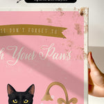 a hand holding a framed canvas print with a black cat and the text "Don't forget to wash your paws" on it.