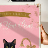 a hand holding a framed canvas print with a black cat and the text "Don't forget to wash your paws" on it.