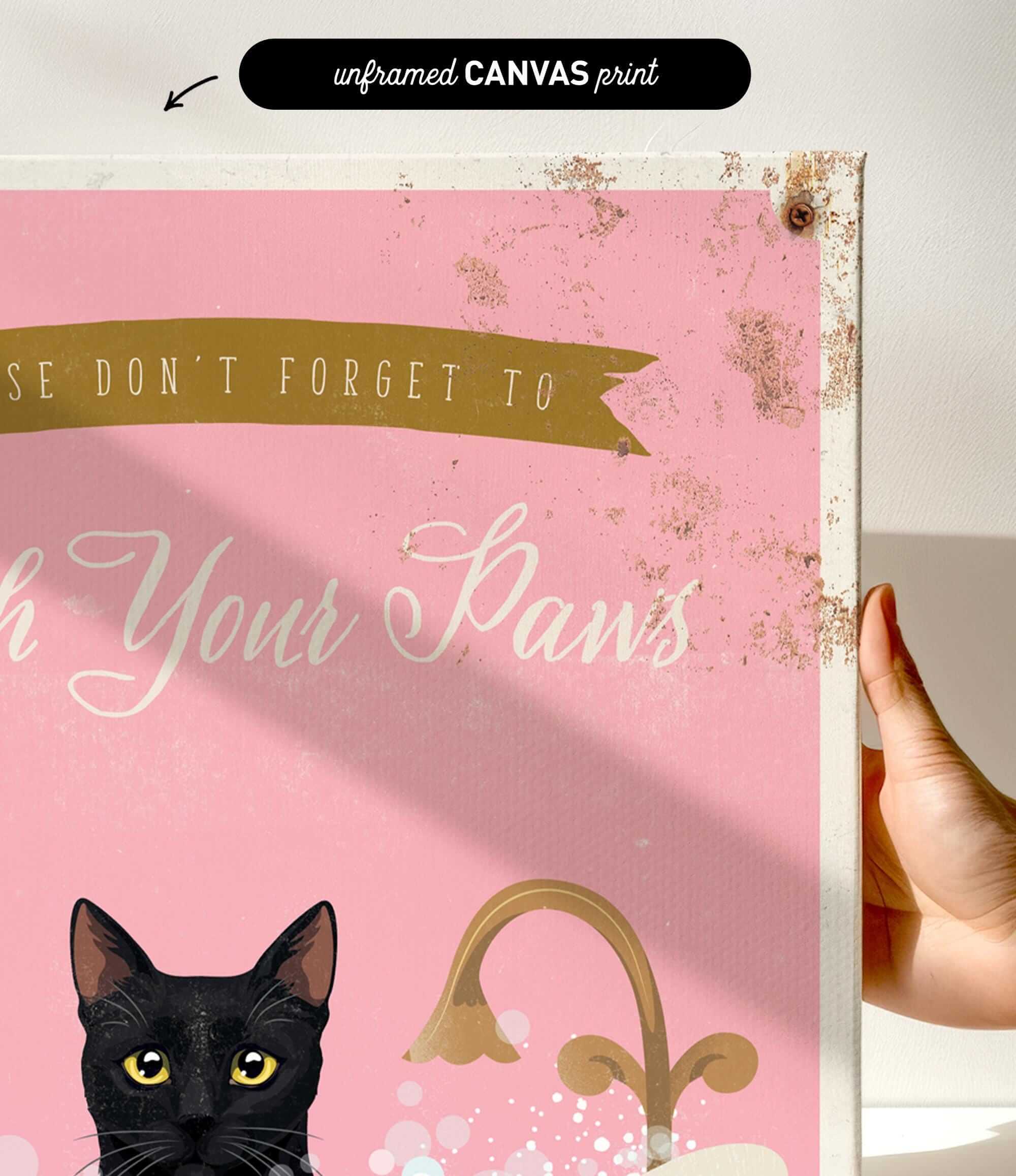 a hand holding a framed canvas print with a black cat and the text "Don't forget to wash your paws" on it.