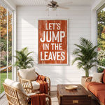 a cozy living room with a large orange sign that reads "LET'S JUMP IN THE LEAVES" hanging on the wall. The room is furnished with wicker chairs and a wooden coffee table, and there are potted plants placed around the space. The overall atmosphere of the room appears warm and inviting, with the sign serving as a focal point.