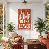 a cozy living room with a large orange sign that reads "LET'S JUMP IN THE LEAVES" hanging on the wall. The room is furnished with wicker chairs and a wooden coffee table, and there are potted plants placed around the space. The overall atmosphere of the room appears warm and inviting, with the sign serving as a focal point.