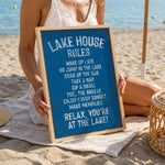 A woman is sitting on a beach holding a blue sign with white text that lists the "Lake House Rules" for a relaxing lakeside experience.