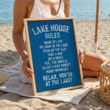 A woman is sitting on a beach holding a blue sign with white text that lists the "Lake House Rules" for a relaxing lakeside experience.