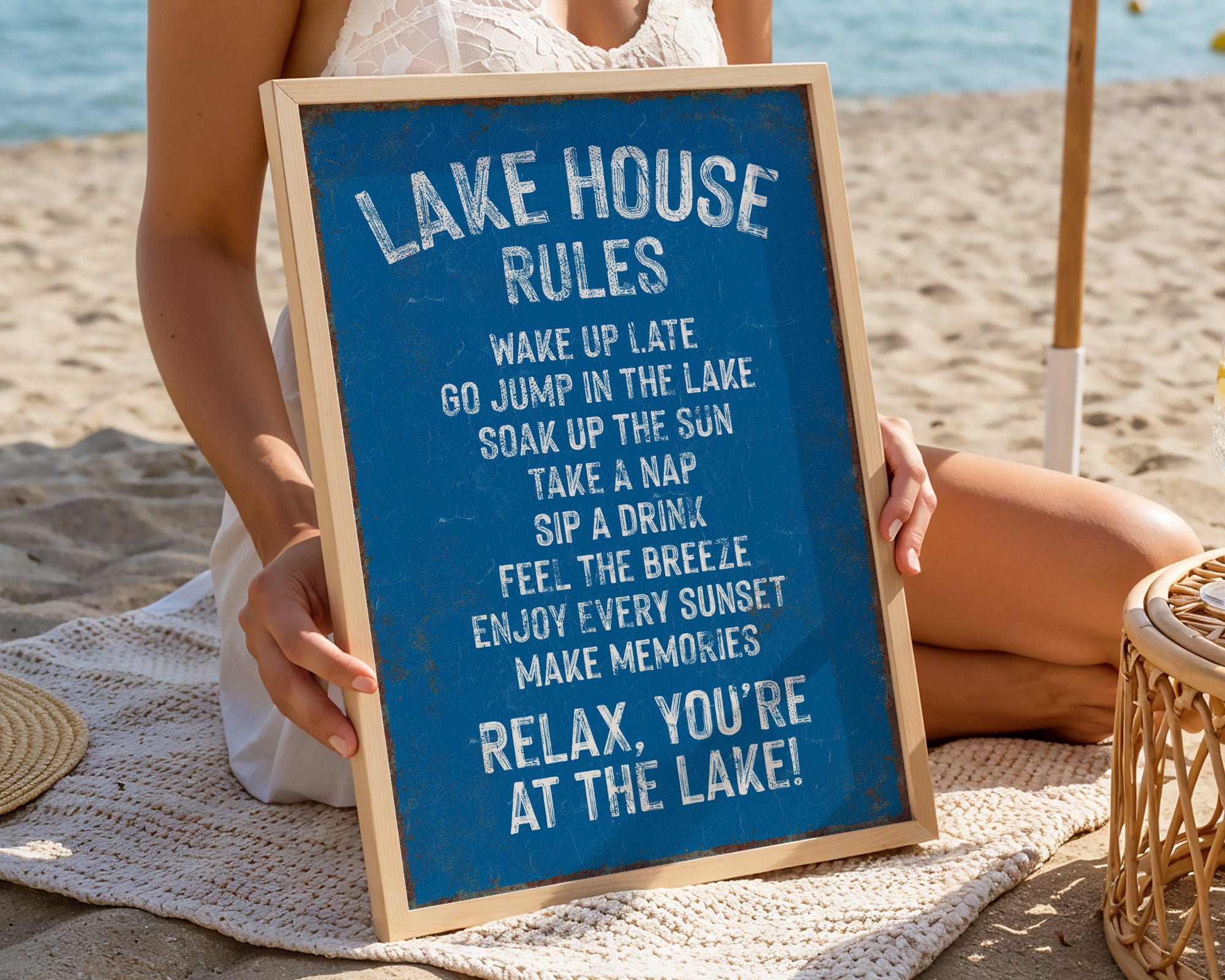 A woman is sitting on a beach holding a blue sign with white text that lists the "Lake House Rules" for a relaxing lakeside experience.