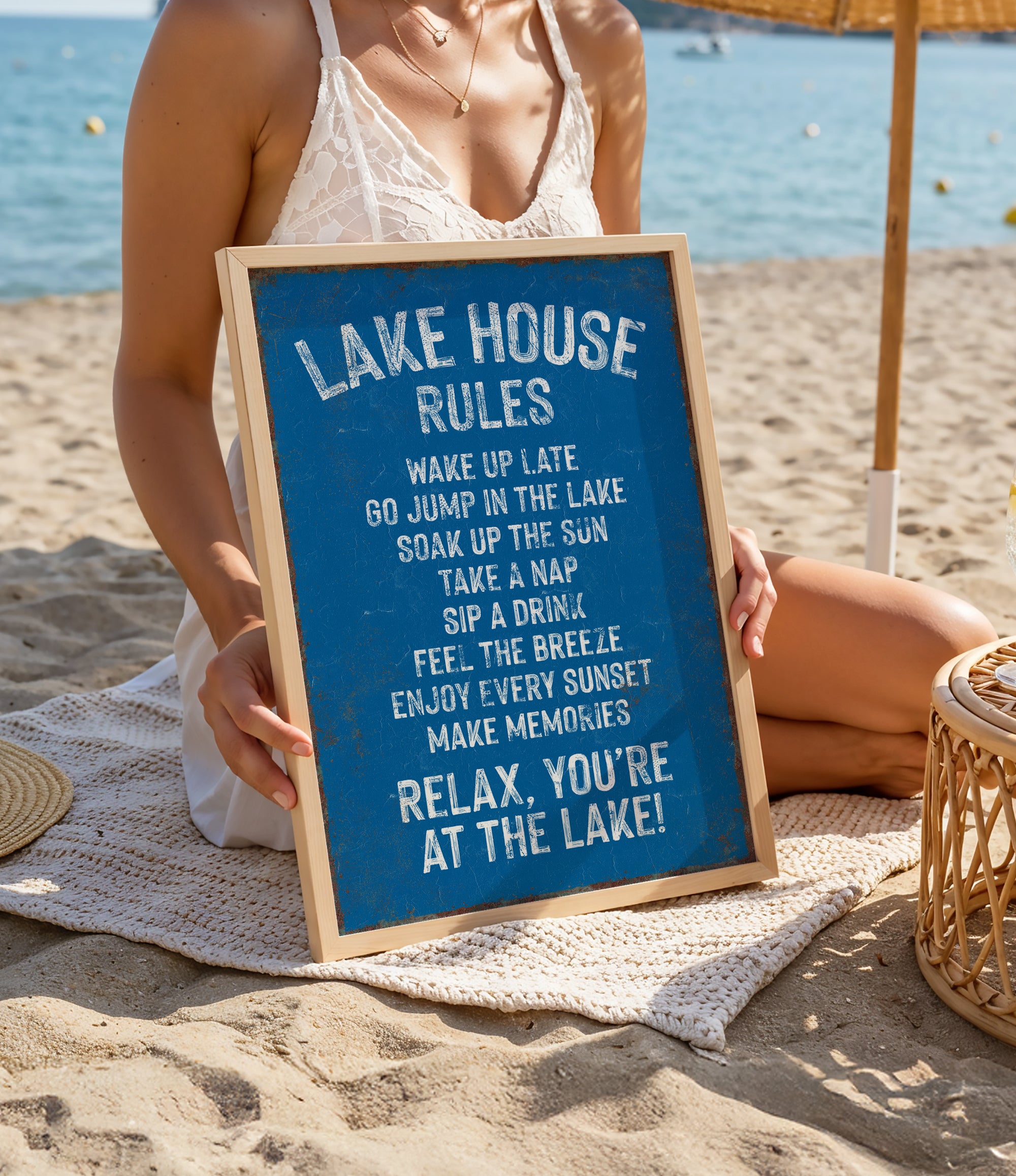A woman is sitting on a beach holding a blue sign with white text that lists the "Lake House Rules" for a relaxing lakeside experience.