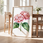a framed watercolor painting of a pink hydrangea flower with green leaves, displayed on a wooden floor in a room with a dining table and chairs.