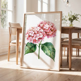 a framed watercolor painting of a pink hydrangea flower with green leaves, displayed on a wooden floor in a room with a dining table and chairs.
