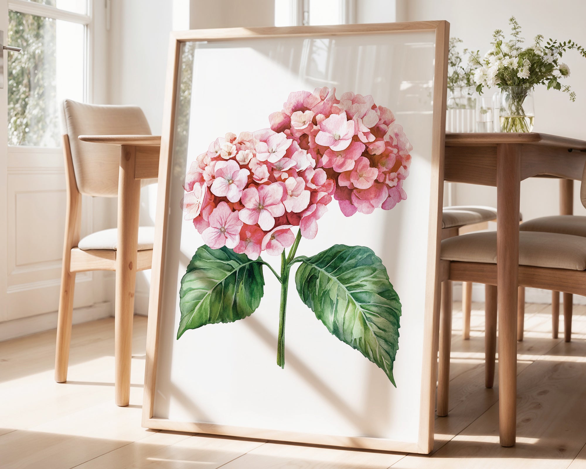 a framed watercolor painting of a pink hydrangea flower with green leaves, displayed on a wooden floor in a room with a dining table and chairs.