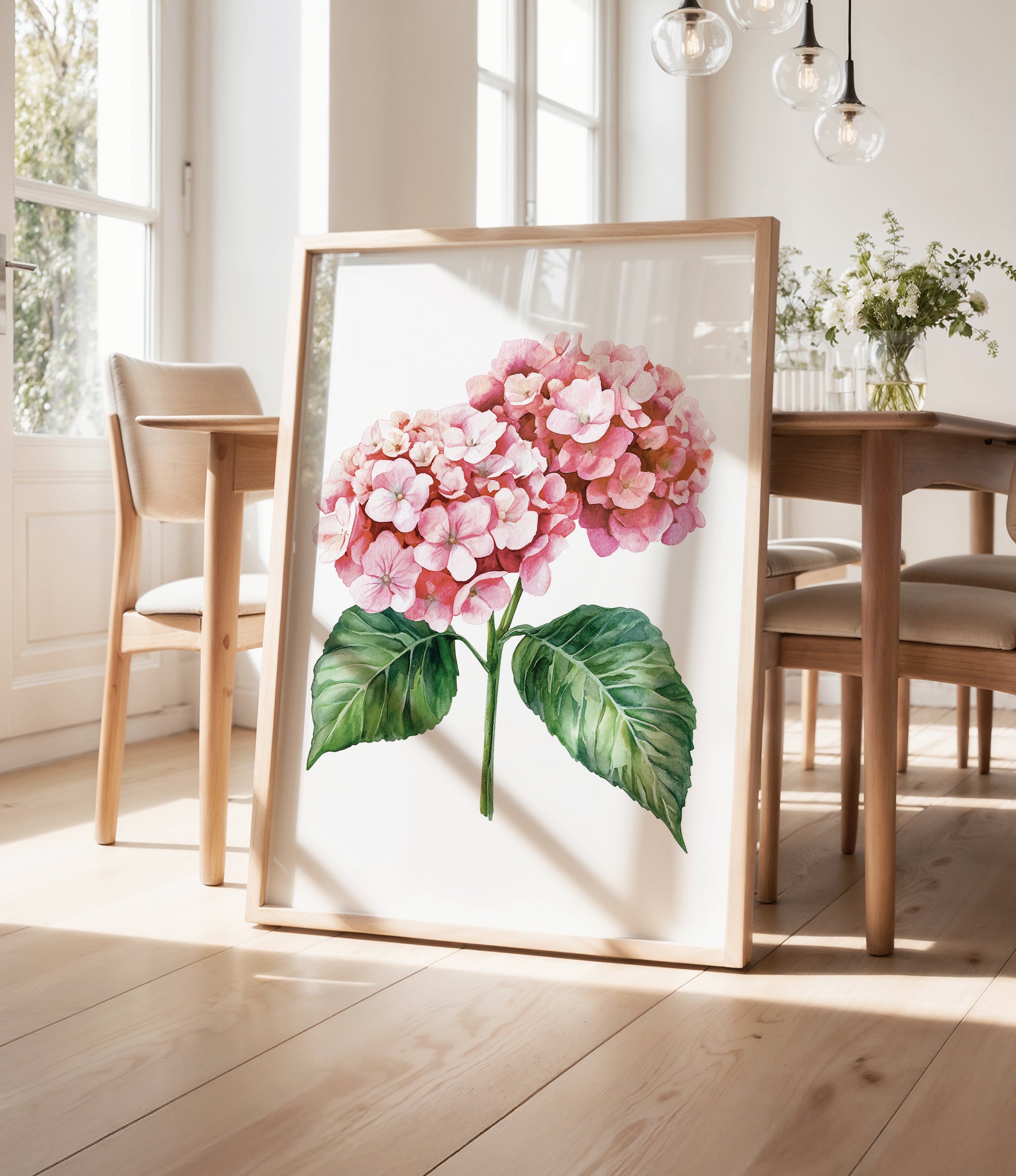 a framed watercolor painting of a pink hydrangea flower with green leaves, displayed on a wooden floor in a room with a dining table and chairs.
