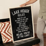 A person is holding up a black sign with white text that reads "LAKESHOUSE RULES" in front of a couch.