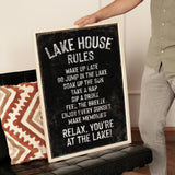 A person is holding up a black sign with white text that reads "LAKESHOUSE RULES" in front of a couch.