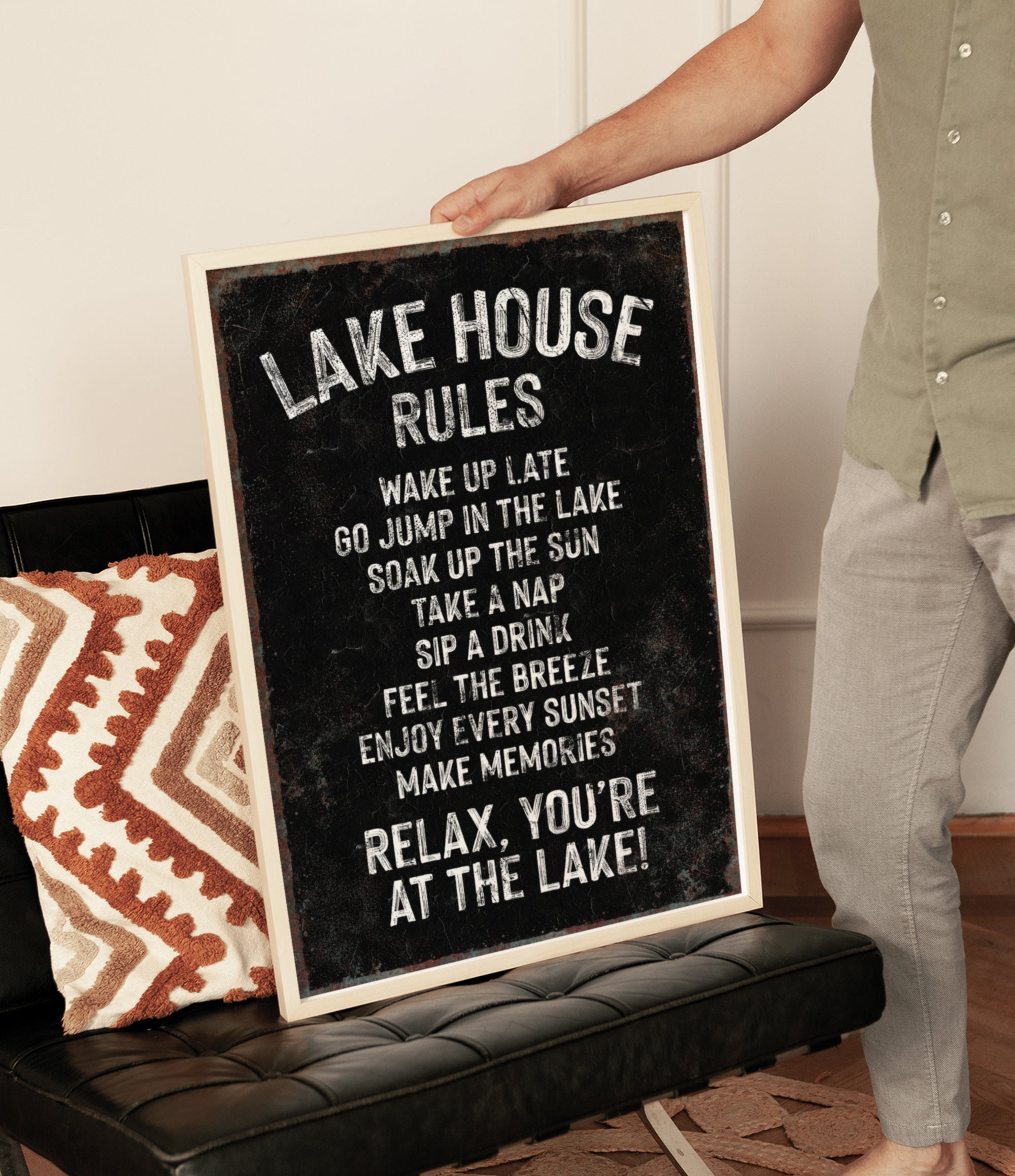 A person is holding up a black sign with white text that reads "LAKESHOUSE RULES" in front of a couch.