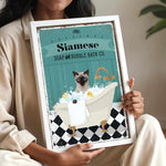 A woman is holding a framed poster with an illustration of a Siamese cat sitting in a bathtub filled with bubbles.