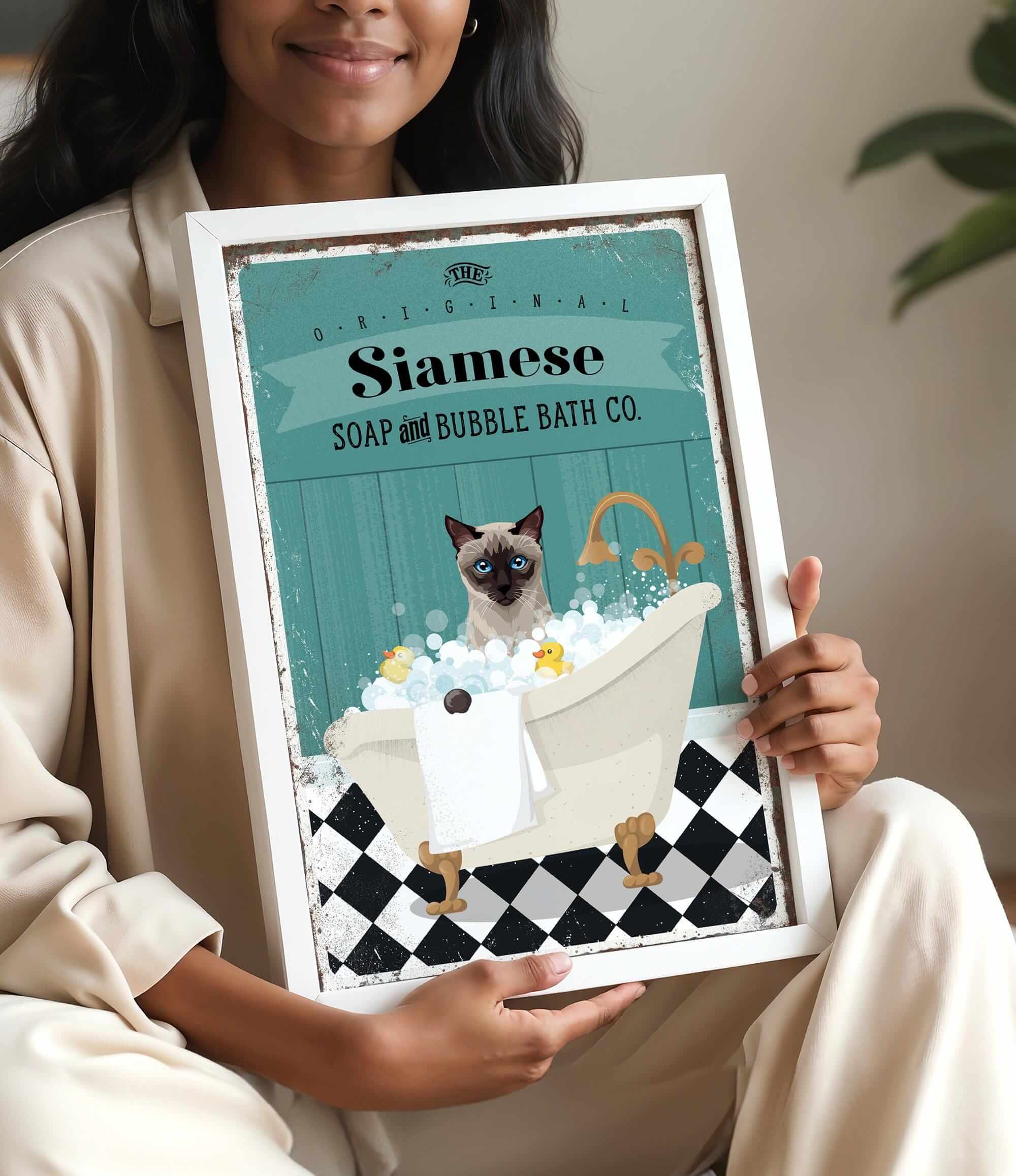 A woman is holding a framed poster with an illustration of a Siamese cat sitting in a bathtub filled with bubbles.