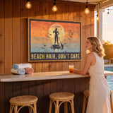 A woman stands at a bar with a framed poster of a paddleboarder and the text "BEACH HAIR, DON'T CARE" above it.