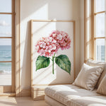 a large framed painting of a pink hydrangea flower with green leaves, placed on a wall near a window overlooking the ocean.