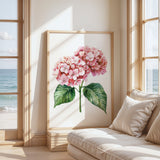 a large framed painting of a pink hydrangea flower with green leaves, placed on a wall near a window overlooking the ocean.